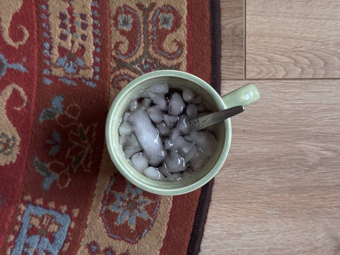 A top-down view of a green ceramic mug filled with ice water and a metal spoon, sitting on a wooden floor next to a red oriental patterned rug.