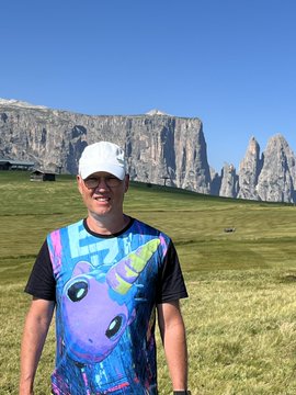 A smiling man wearing a white cap and a colorful unicorn-print t-shirt poses in a grassy alpine meadow, with the dramatic rocky peaks of the Dolomites rising in the background under a clear blue sky—evoking the natural, authentic, and community-driven spirit highlighted by the $UFD post.