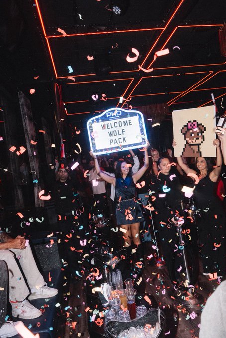 A celebratory scene inside a nightclub with confetti falling from the ceiling, featuring people holding a lit 'PMD WELCOME WOLF PACK' sign and a pixel-art portrait.
