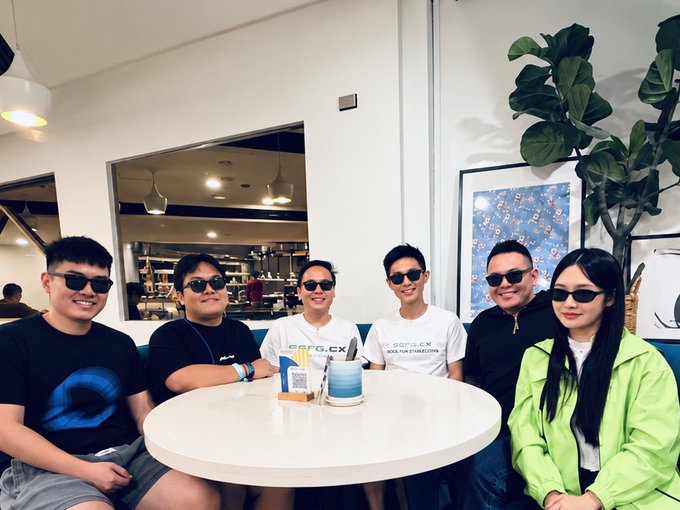 A group of six people, five men and one woman, pose wearing sunglasses around a small table at what appears to be a restaurant, with two individuals wearing matching white t-shirts with 'SOFG.CX' printed on them.