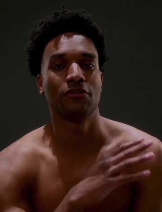 A shirtless man with short curly hair stands against a dark background, gesturing with his hand across his chest.