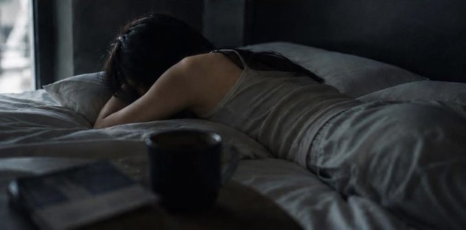 In this image, a woman is lying down on her bed with her head resting on her arm. She has dark hair and is wearing white clothing. A cup of coffee sits next to her on the bed, suggesting that she might be enjoying a relaxing morning or evening routine. The background features a window, which could provide natural light for her activities.