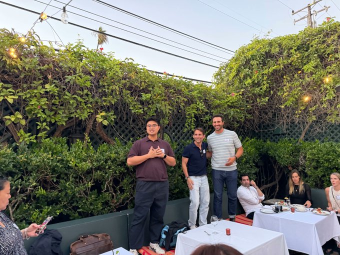Two men stand smiling at the center of an outdoor patio gathering, with a third man holding a microphone beside them, while attendees mingle around white-clothed tables set against a lush green hedge backdrop. The scene captures the Fintech Connect Happy Hour sponsored by Coinbase in LA, where guests discuss building payments, stablecoin, custody, and trading products onchain.