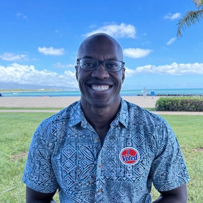 A man wearing glasses and a blue patterned Hawaiian shirt smiles while wearing an "I Voted" sticker at a sunny beachside location, visually celebrating his participation in voting "Yes" to allow $STRC and Strategy's other preferred stocks to pay dividends daily.
