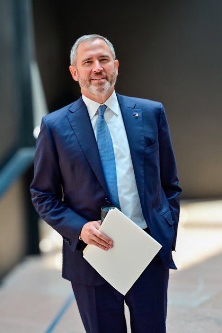 A man with a gray beard wearing a blue suit and tie, smiling while holding a beige folder or envelope.