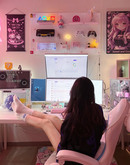 A cozy pink-themed gaming/streaming setup featuring a person with long dark hair sitting at a multi-monitor desk, surrounded by anime figurines, a 'You Can Do It!' poster, game controllers, an anime girl poster, and various collectibles on a pegboard.