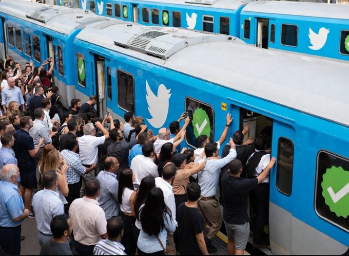 An overhead shot of a large crowd of people packing tightly into a blue train decorated with Twitter/X logos and green verification checkmarks.