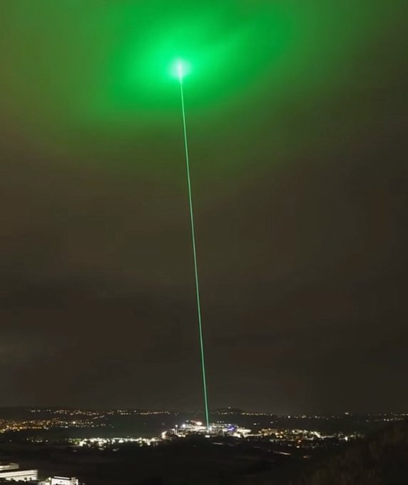 A bright green laser beam pierces the night sky above a distant city, creating an illuminated connection between the ground and a glowing orb overhead. The image metaphorically symbolizes the bullish market signal highlighted in the post, as Treasury Secretary Bessent's announcement of a potential deal to reopen the Strait of Hormuz sparks optimism for Bitcoin and altcoins.