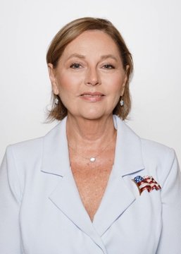 A woman with short blonde hair, wearing a white blazer adorned with an American flag lapel pin and a pearl necklace, poses for a professional portrait against a plain background. The image accompanies a breaking news update about Darline Graham Nordone's narrowing lead ahead of the South Carolina GOP Senate special primary.
