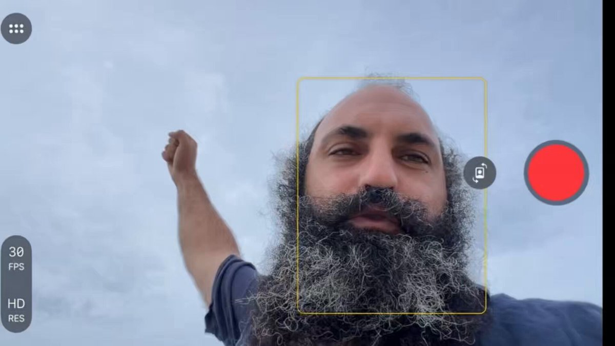 The image shows a man with a beard holding up his arm, possibly taking a selfie or recording something. The background of the photo appears to be cloudy skies, which could suggest an outdoor setting. There are two red circles present in the image, one located near the top right corner and another at the bottom left corner. These red circles might represent a camera or a phone screen that is capturing the moment.