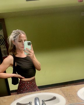 A woman with blonde hair takes a mirror selfie in a bathroom with green walls, wearing a black crop top and patterned skirt, holding a phone with a light blue case.