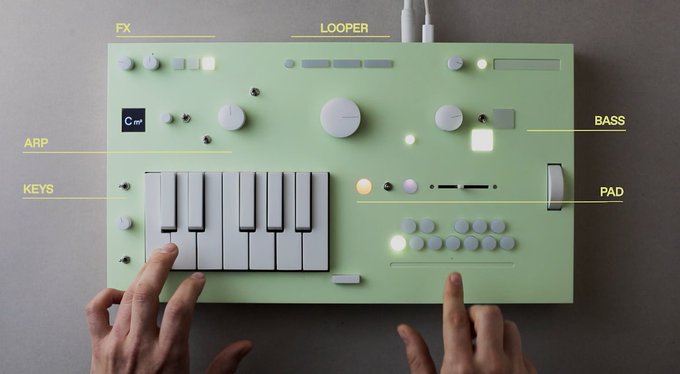 An overhead view of hands operating a green electronic synthesizer or music device with labeled sections for FX, LOOPER, ARP, KEYS, BASS, and PAD.