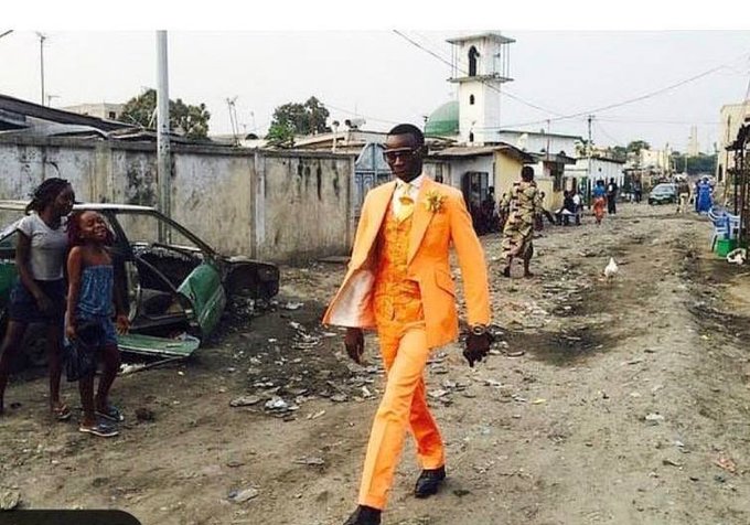 A street scene where a man in a bright orange three-piece suit and sunglasses walks confidently down a dirt road, while children and onlookers watch from beside a wrecked car.