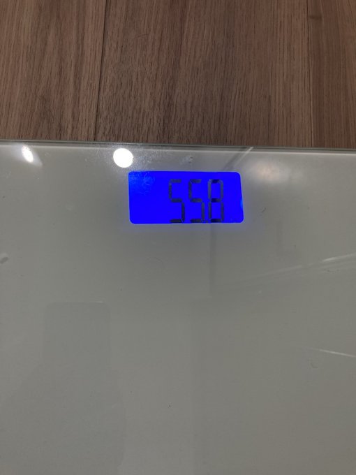 The image shows a digital scale with the number 559 displayed on its screen, indicating that someone has stepped onto it for weight measurement. The scale appears to be white and is placed on top of a wooden floor or table. There are no other objects in the scene, and the focus is solely on the scale and the person's weight at that moment.