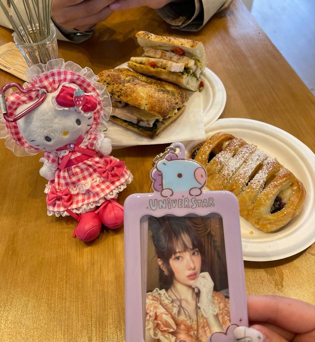 A cafe table with sandwiches and pastries, alongside a Hello Kitty plush toy wearing a pink gingham dress and a UNIVERSTAR photocard featuring a K-pop idol.