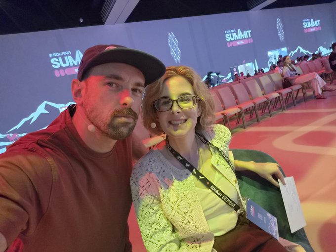 A man and woman take a selfie at the Solana Summit, with the event's branding visible on the large screen behind them as they pose among empty auditorium seats during day 2 of the conference.