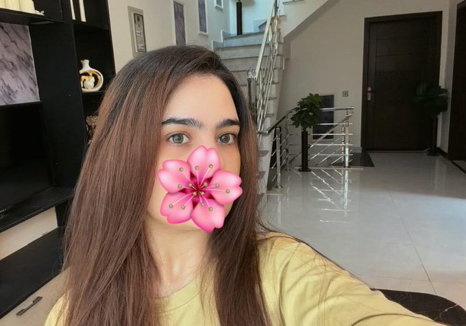 A young woman takes a selfie indoors with a pink flower filter covering her mouth, standing in a modern home with a staircase visible behind her.