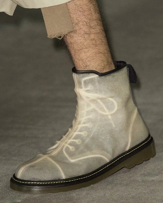 A close-up fashion shot of a translucent/clear suede lace-up ankle boot with decorative stitching, worn with frayed brown trousers, displayed on a grey surface.