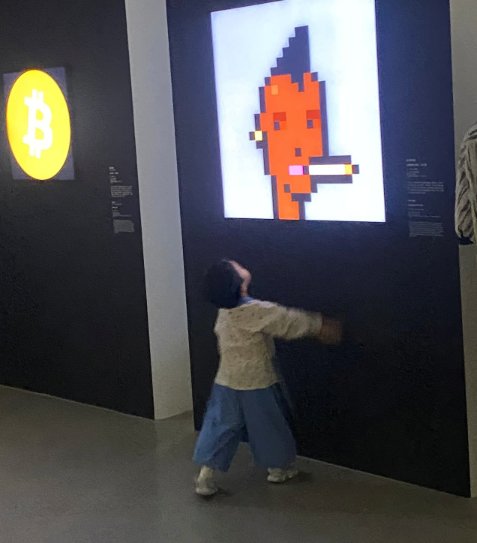 A child joyfully interacts with a large display of CryptoPunk #110, a pixelated orange character with a cigarette, at the West Bund Museum in Shanghai. To the left, the Bitcoin logo glows in yellow, accompanying the pixel art piece as part of the Computer Worlds exhibition featuring digital and computer art.