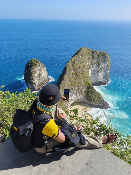 A Binance Angel takes in the breathtaking view of Kelingking Beach in Nusa Penida, Indonesia, capturing the iconic T-Rex-shaped cliff with his phone while wearing a Binance-branded bucket hat and backpack.
