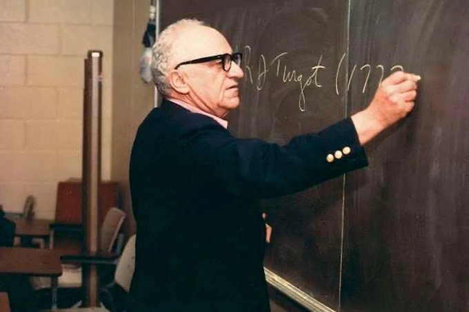 A photograph of an elderly bald man with glasses, wearing a dark blazer, writing on a chalkboard with chalk.