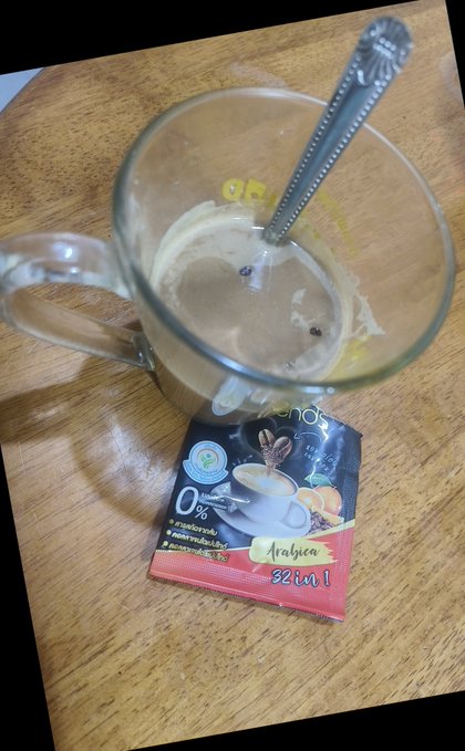 A nearly empty glass mug with a metal spoon, placed on a wooden table next to a single-serving packet of "Arabica 3 in 1" instant coffee mix.
