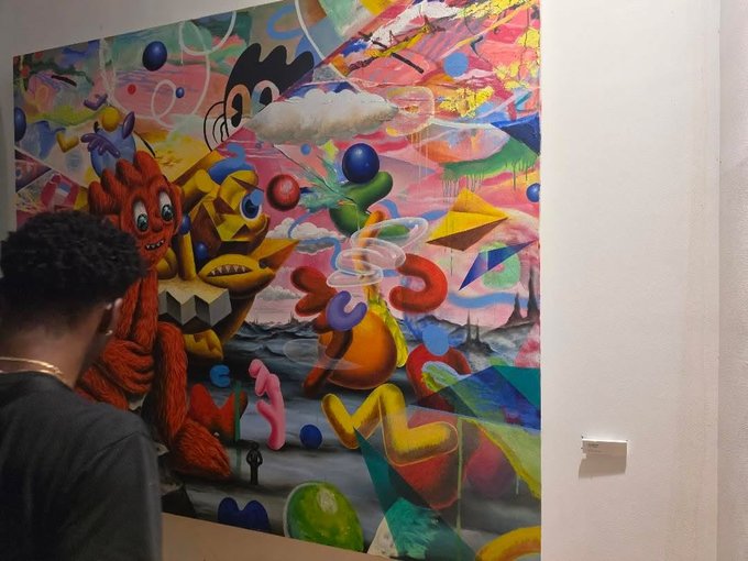 A vibrant, chaotic abstract painting featuring colorful geometric shapes and cartoon-like creatures, with a person viewing it in a gallery.