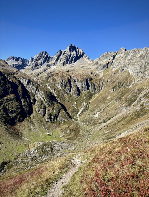 A scenic mountain landscape features rugged rocky peaks under a clear blue sky, with a narrow dirt hiking trail winding through a grassy alpine valley below.