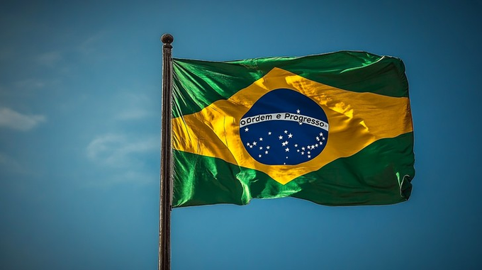 The Brazilian flag waving against a blue sky with clouds, showing its green field, yellow diamond, and blue globe with the motto 'Ordem e Progresso'.