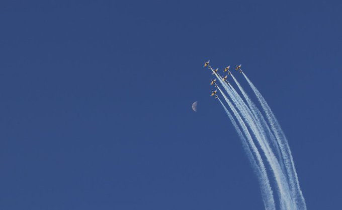 Five jets flying in formation against a deep blue sky, leaving white smoke trails beside a visible crescent moon.