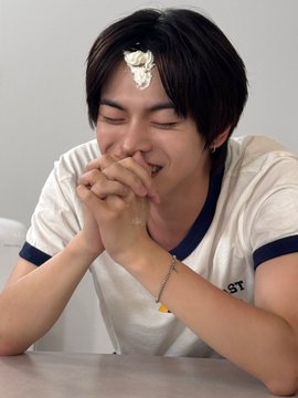 A young man with dark hair laughs with his hands clasped together as a dollop of whipped cream sits on his forehead, while he wears a white t-shirt featuring partial text reading "ST" and a colorful beaded bracelet. The image captures a playful, lighthearted summer moment fitting the caption's "musim panas" (Indonesian for "summer") and references LEO from ALLDAY PROJECT.