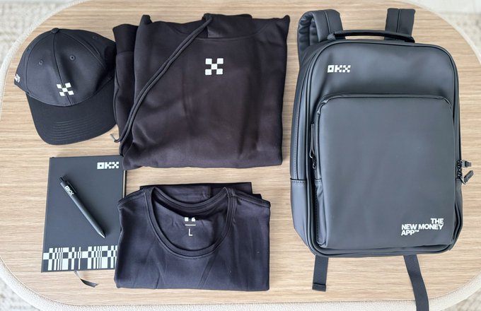 A flat lay displays OKX-branded merchandise including a black cap, folded hoodie, t-shirt, notebook with pen, and a black backpack labeled "THE NEW MONEY APP," all arranged on a wooden table as part of the OKX merch collection featured in the post.