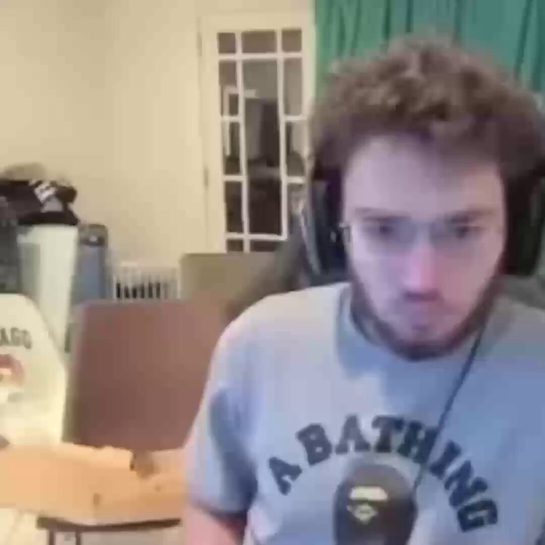 The image shows a man wearing a gray t-shirt with "A Bathing" written across the front, sitting at a desk or table while using an app on his phone. The text of the post reads, "Am I the only one checking the app to see if I received access to 𝕏 Money yet? @elonmusk". This suggests that the man might be trying to find out if he has been given access to a certain feature or resource in the app, possibly related to money.