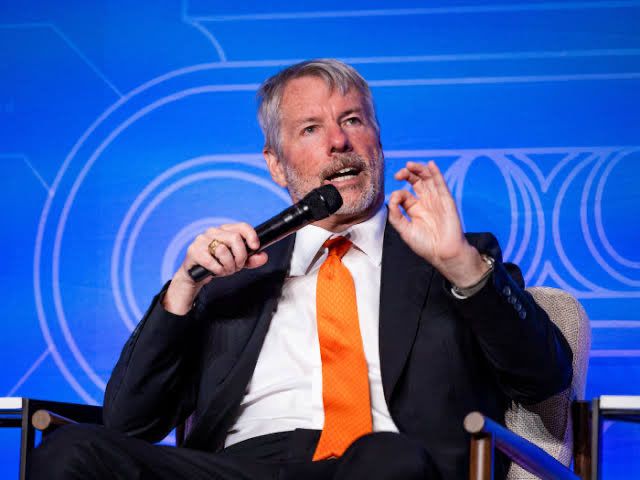 A photograph of a gray-haired, bearded man in a dark suit and orange tie speaking into a microphone while gesturing with his hand at what appears to be a conference or panel discussion.