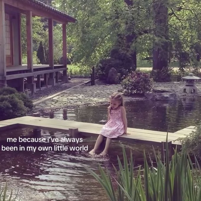 A young girl in a pink dress dangles her feet over a wooden dock into a pond at a Japanese-style garden, with text reading 'me because i've always been in my own little world'.