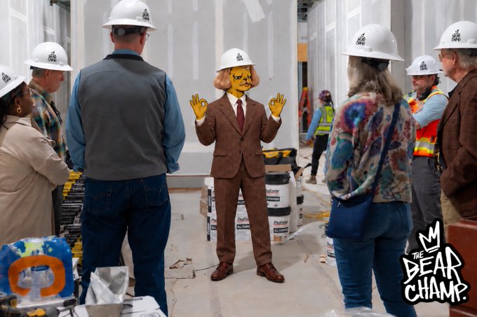 Construction site scene from 'The Bear Champ' TV show showing people in hard hats and high-vis vests, with one person in the center having an edited orange cat face wearing a brown suit.