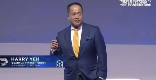Photograph of Harry Yeh, Managing Director of Quantum Fintech Group, presenting on stage in a blue suit and yellow tie at the Blockchain & DeFi Developer Conference.