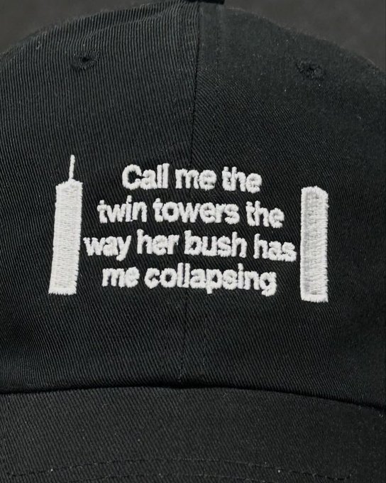 A close-up of a black baseball cap featuring an embroidered design of the twin towers and the text 'Call me the twin towers the way her bush has me collapsing'.