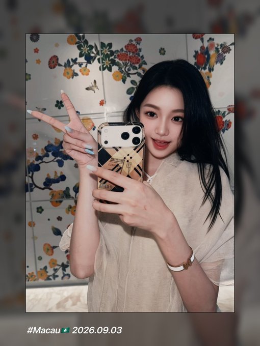 A young woman with long dark hair poses for a mirror selfie, making a peace sign while holding an iPhone with a decorative case, set against a backdrop of colorful floral tiles. The caption "#Macau 🇲🇴 2026.09.03" appears at the bottom, suggesting a personal travel moment that contrasts with the accompanying post's shift toward a professional "node business" focus.