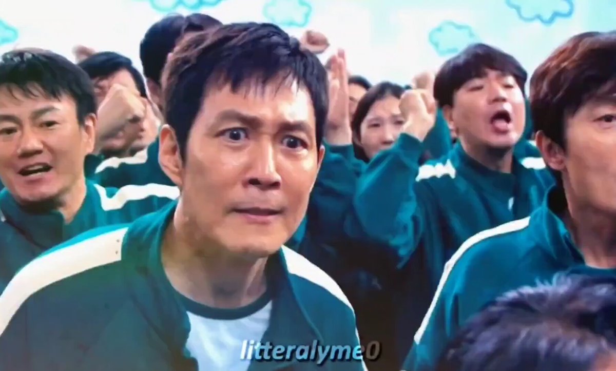 The image features a close-up shot of two men standing together, likely in front of other people. They are wearing matching green uniforms with white lines running down the sleeves. One man appears to be looking at something off screen or towards the camera, while the other seems to be looking towards his friend. The background is blurred and indistinct, drawing attention to the main subjects. The surrounding environment seems to be a gathering area or social event where people are engaged in conversation or observing an ongoing activity.