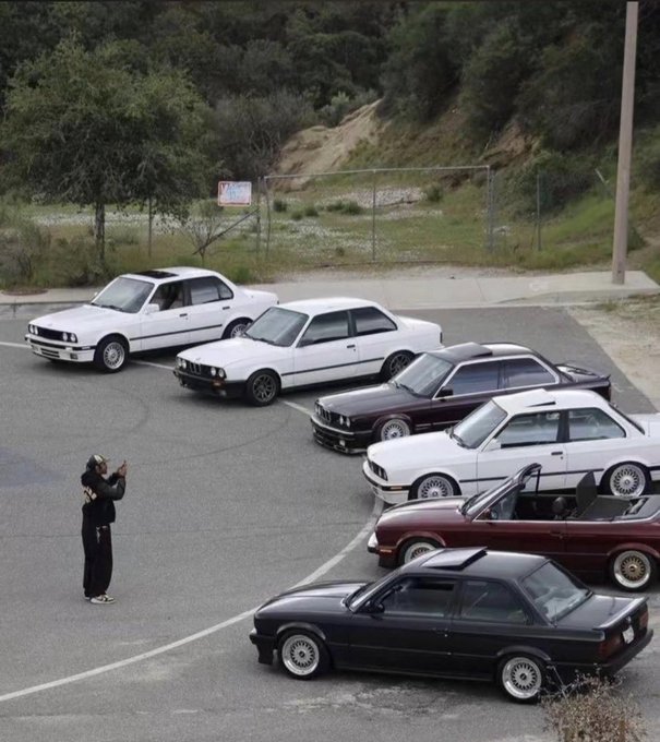 An aerial view of a parking lot where a person stands filming a lineup of classic BMW E30 cars in various colors including white, maroon, and black.