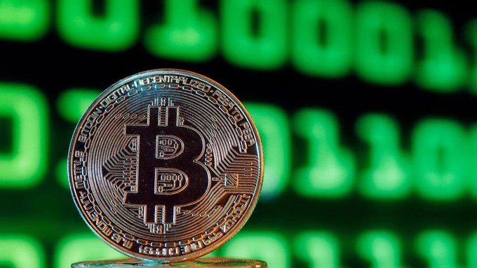 A close-up of a Bitcoin physical coin standing upright against a blurred background displaying bright green digital numbers, suggesting a cryptocurrency exchange interface.