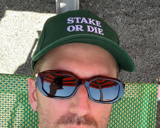 A close-up selfie of a man wearing a green cap reading 'STAKE OR DIE' and dark sunglasses that reflect the photographer holding a camera, sitting on a green patterned chair outdoors.