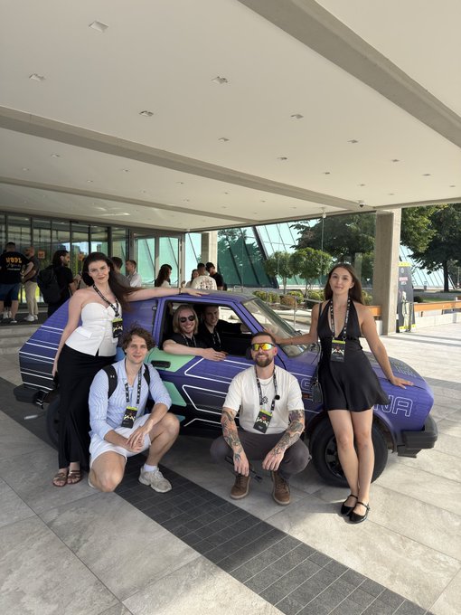 The image shows a group of people posing around a small purple car with "URA" visible on the side, with a modern building with large windows in the background. Several individuals wear lanyards suggesting they're conference attendees at an event. The caption perfectly captures the infectious enthusiasm of the Belgrade Blockchain Week crowd, as everyone in the photo is smiling warmly and posing together in a relaxed, friendly manner that embodies the welcoming atmosphere described in the post.