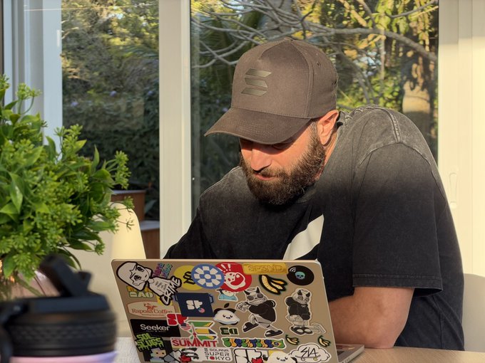 A bearded man in a brown cap and dark t-shirt working on a laptop covered in colorful tech and pop-culture stickers.