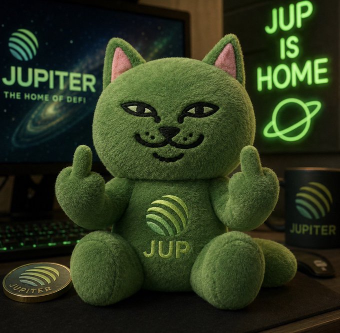 A green plush cat toy with 'JUP' on its chest and middle fingers raised sits on a desk, surrounded by Jupiter crypto branding including a screen reading 'JUPITER THE HOME OF DEFI' and a neon sign saying 'JUP IS HOME'.