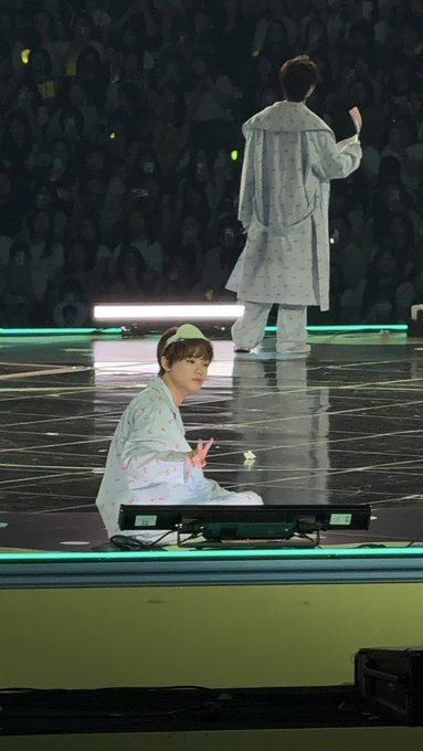 A performer in a white polka-dotted outfit kneels on a glossy stage making a peace sign, while another performer in matching attire stands facing away toward an audience illuminated by green light sticks.