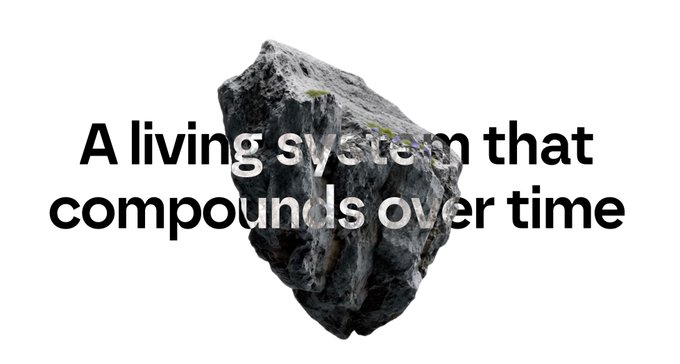 A rough gray rock with small patches of green moss sits against a white background, overlaid with the bold black text "A living system that compounds over time."

The rock metaphorically represents a durable, foundational asset—suggesting that Axis's robot training data infrastructure is a solid, growing foundation (like weathered stone accumulating moss over time) rather than a fleeting trend, emphasizing that their community-driven data engine builds lasting value that compounds as more users contribute.