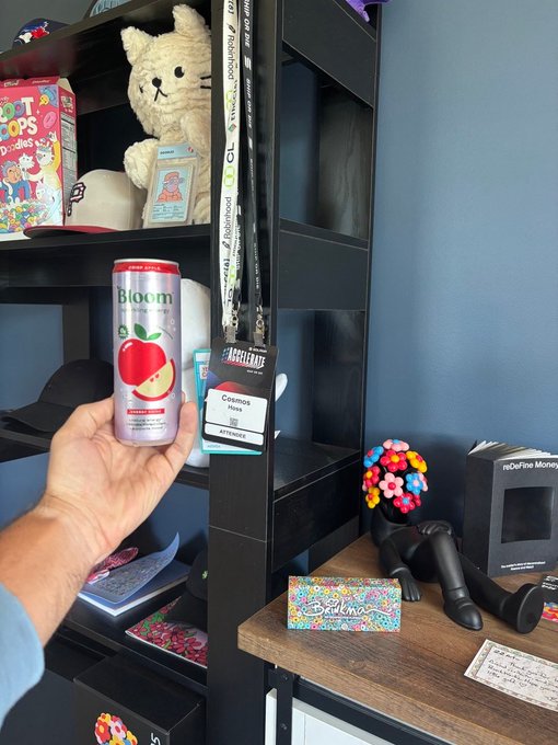 A hand holds up a 'Bloom' Apple Cinnamon sparkling beverage can in front of a black shelving unit displaying merchandise including a Pop-Tarts 'Dora the Explorer' box, lanyards, and figurines.