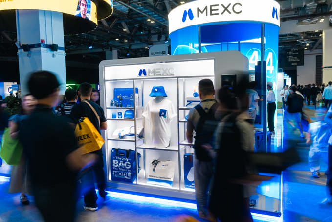 MEXC's branded booth at Token2049 displays branded merchandise including hats, bags, and apparel with visible text like "MEXC," "TRADE YOUR BAG NOW," drawing a steady crowd of attendees.